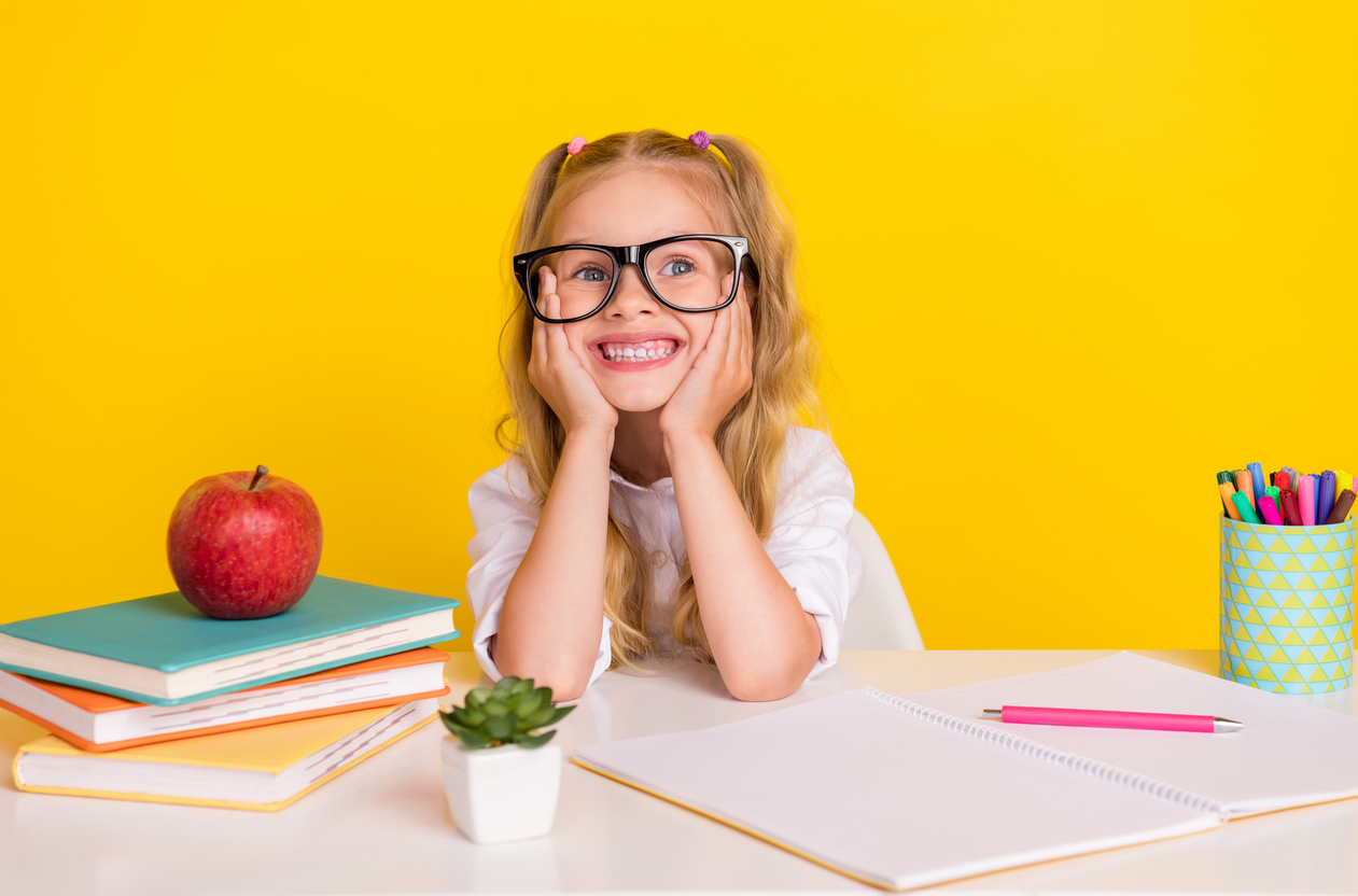 Photo of adorable dreamy little lady dressed white shirt eyewear sitting table arms cheeks looking empty space isolated yellow color background